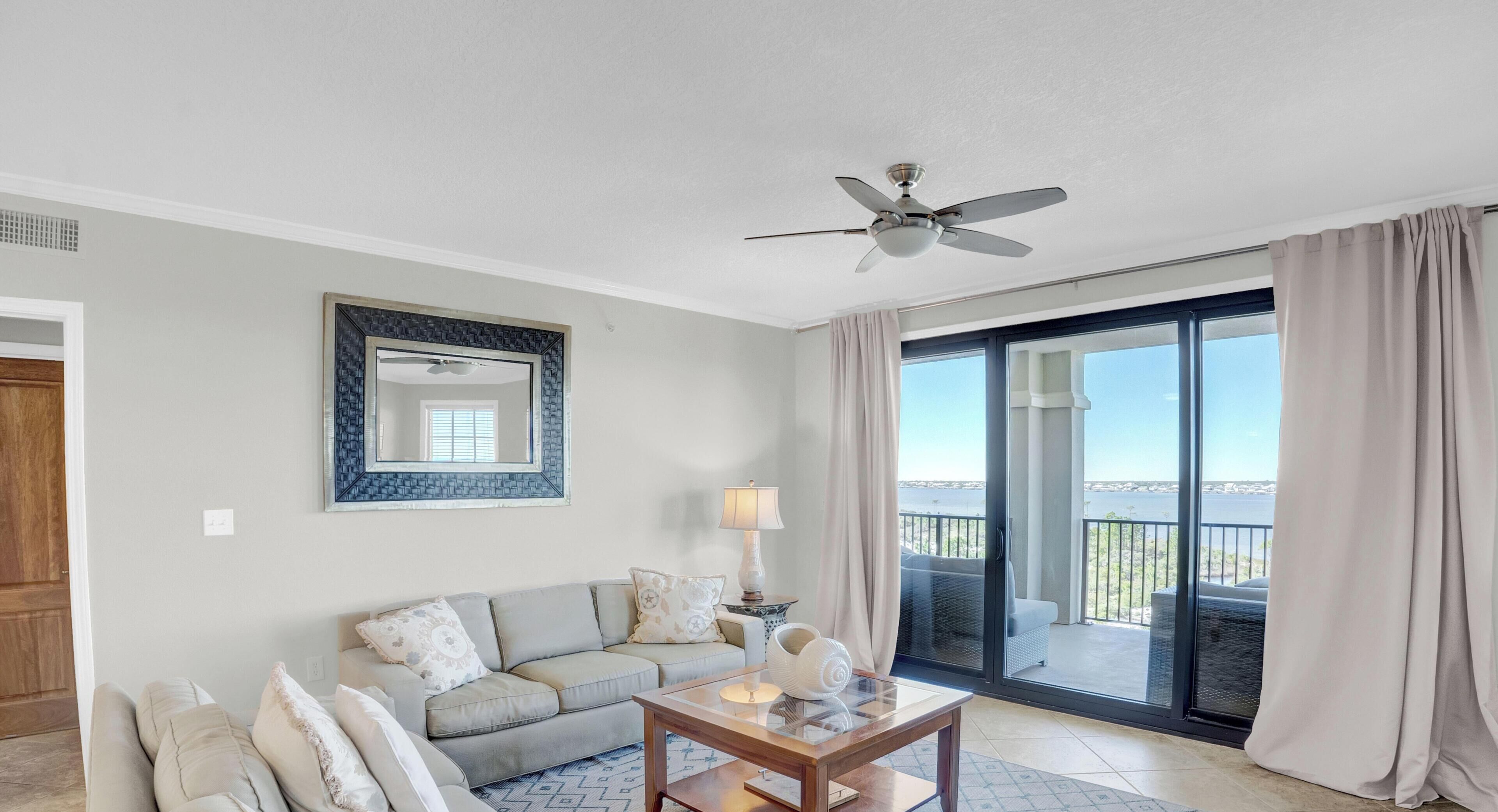 A living room with a sofa, a coffee table, a lamp, a ceiling fan, a large mirror, and a sliding glass door leading to a balcony with a view.