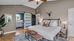 A bedroom with a large bed, a bench, a ceiling fan, and a view into the kitchen. - New! Wildwood Manor ~ Salt Water Hot Tub ~ Dog Friendly ~ Fire Pit (Bend)