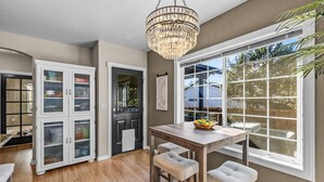 A kitchen with a wooden table, two stools, a chandelier, and a window with a view of the outside. - New! Wildwood Manor ~ Salt Water Hot Tub ~ Dog Friendly ~ Fire Pit (Bend)