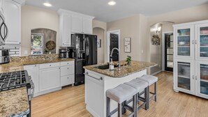 A modern kitchen with white cabinetry, granite countertops, a central island with bar stools, and a stainless steel refrigerator. - New! Wildwood Manor ~ Salt Water Hot Tub ~ Dog Friendly ~ Fire Pit (Bend)