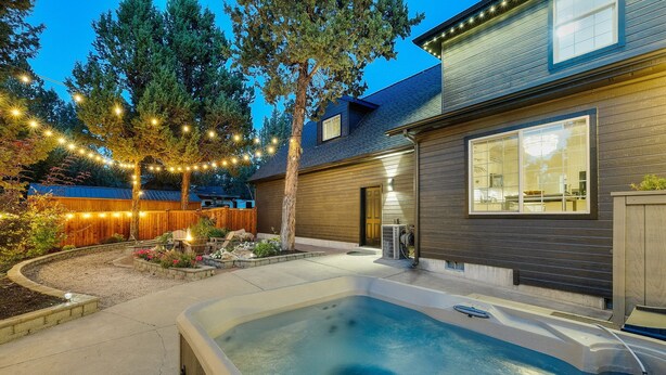 A backyard with a hot tub, string lights, a wooden fence, and a house with large windows. - New! Wildwood Manor ~ Salt Water Hot Tub ~ Dog Friendly ~ Fire Pit (Bend)