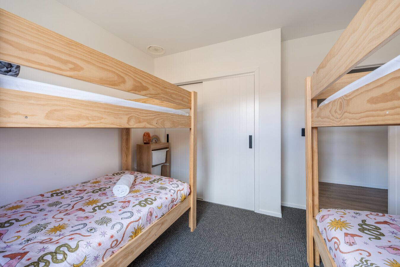 Una habitación con literas, una puerta blanca y un pequeño estante de madera con objetos.