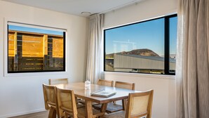 Un comedor con mesa y sillas de madera, ventanas grandes con cortinas y vistas a un edificio y paisaje.