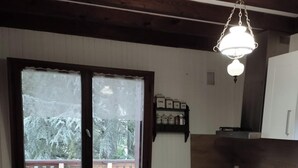 Een eethoek met een ronde tafel, stoelen, een raam met uitzicht op bomen, een keuken met kasten en apparatuur, en een hangende lamp.
