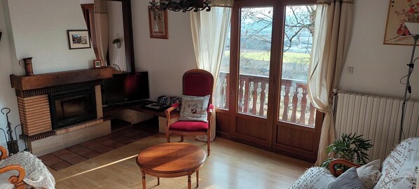Un salon chaleureux avec une cheminée, un fauteuil rouge, une table basse en bois et une vue à travers une porte vitrée.