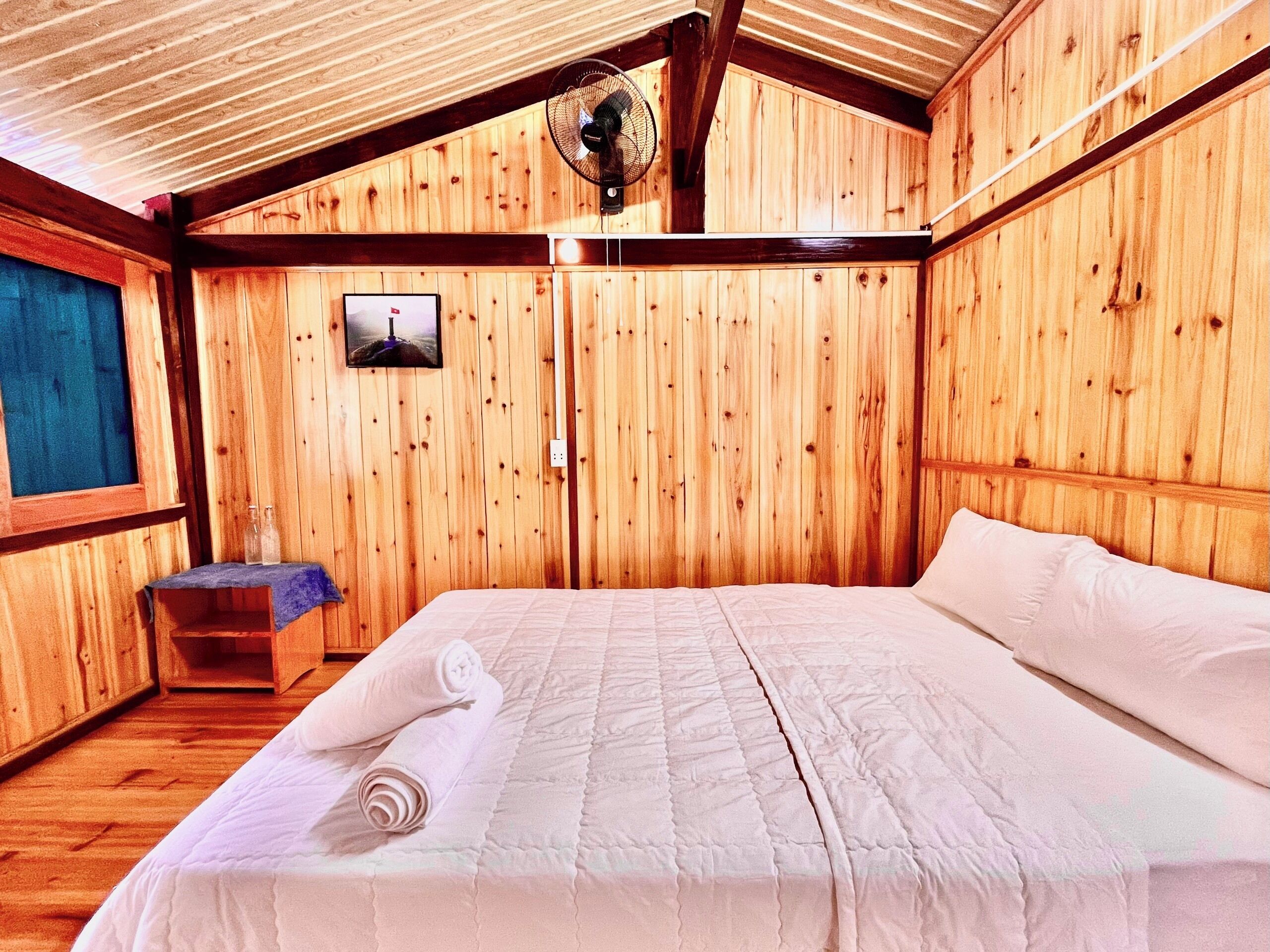 Ein Zimmer mit Holzboden, einem Bett, einem Ventilator und einem kleinen Tisch mit einem Bild an der Wand.