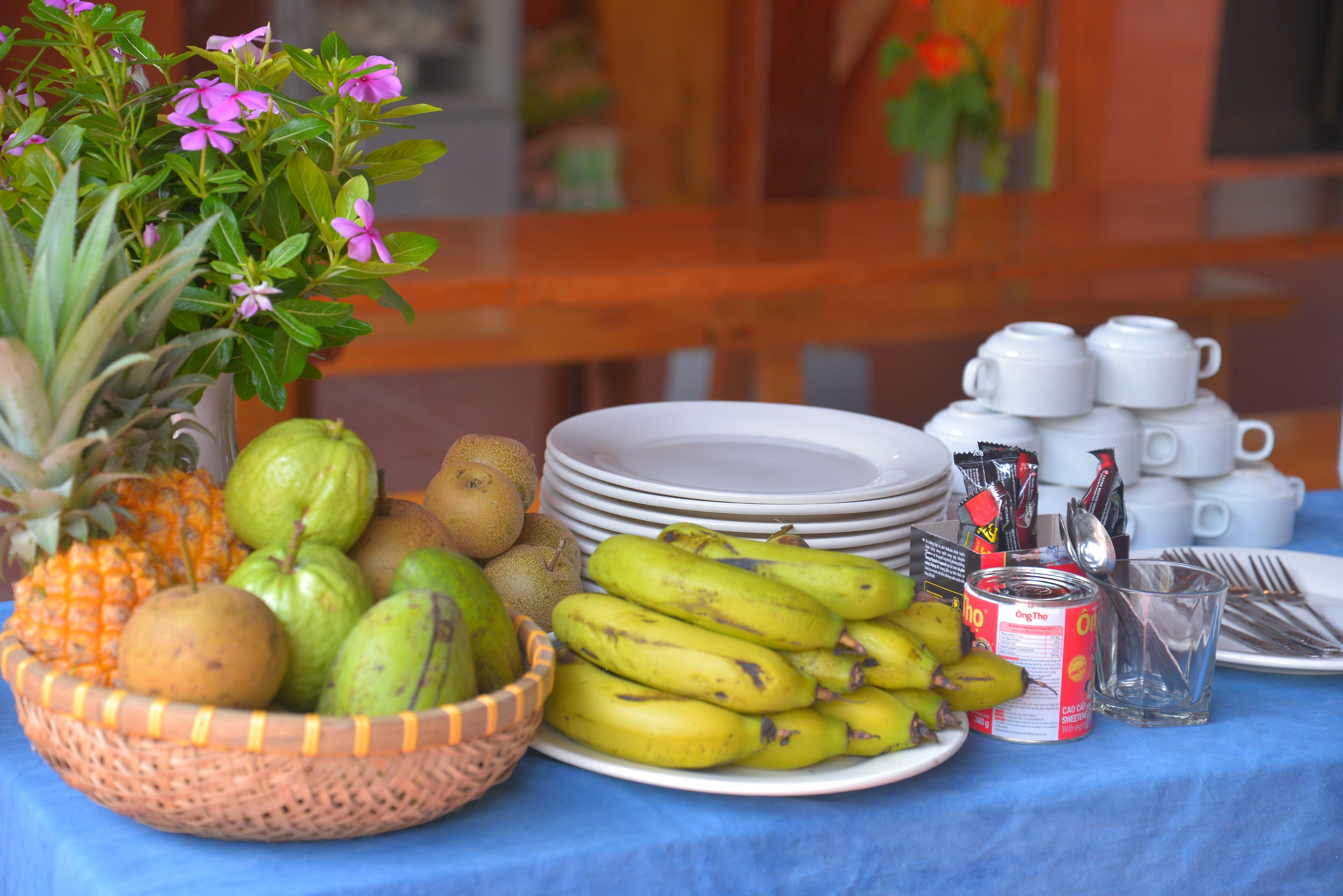 A table with a fruit basket, plates, a can, and a stack of cups.