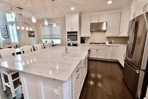 A kitchen with white cabinets, a large island with a sink, stainless steel appliances, pendant lighting, and a dining area with chairs.