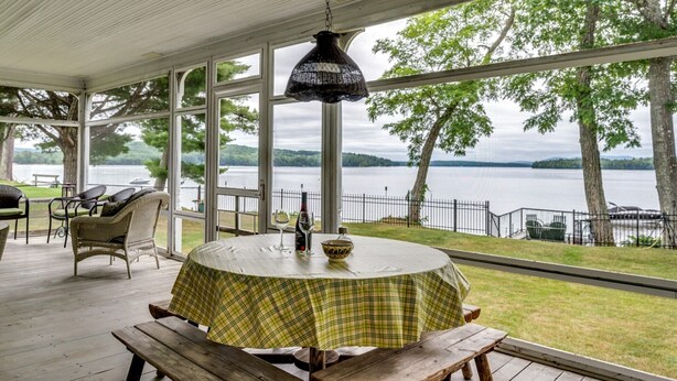 A covered patio with a table and chairs, overlooking a lake with trees and a dock. - Winnipesaukee waterfront escape with private dock and guest cottage (Tuftonboro)