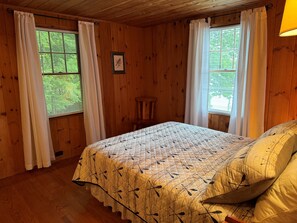 A wooden-paneled room with a bed, two windows with curtains, a framed picture on the wall, and a small stool. - Charming Pet Friendly Lakeside Escape with Cozy Comfort and a Private Dock (Moultonborough)