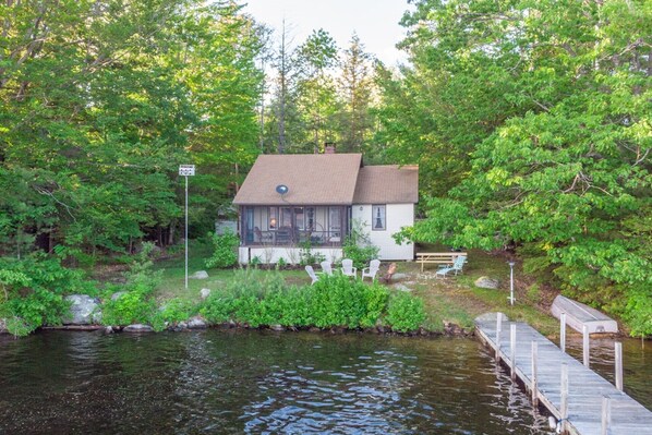 A small house with a dock extending into a calm body of water, surrounded by dense greenery. - Charming Pet Friendly Lakeside Escape with Cozy Comfort and a Private Dock (Moultonborough)
