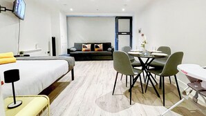A modern hotel room with a bed, a dining table with chairs, a sofa, and a TV mounted on the wall.
