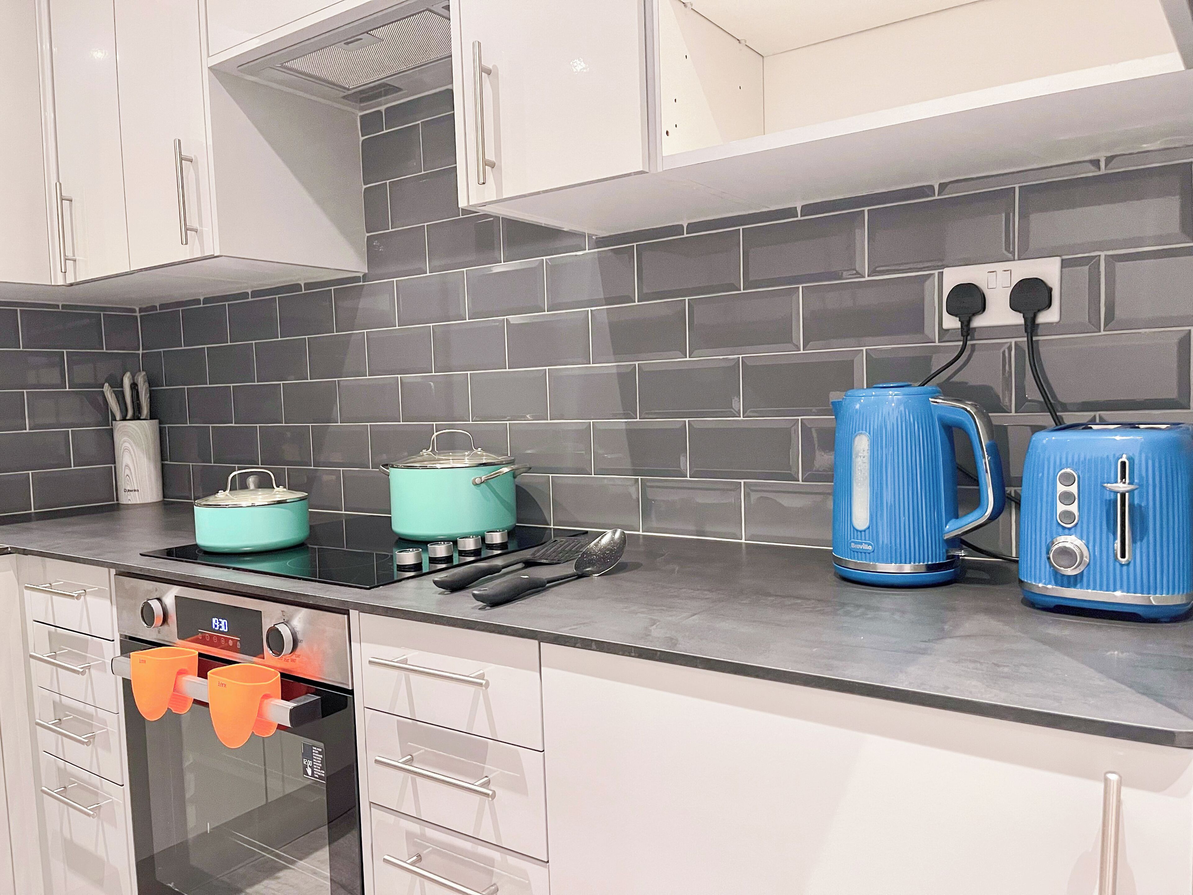 A modern kitchen with grey tiled backsplash, white cabinets, a stove, and two blue kettles.