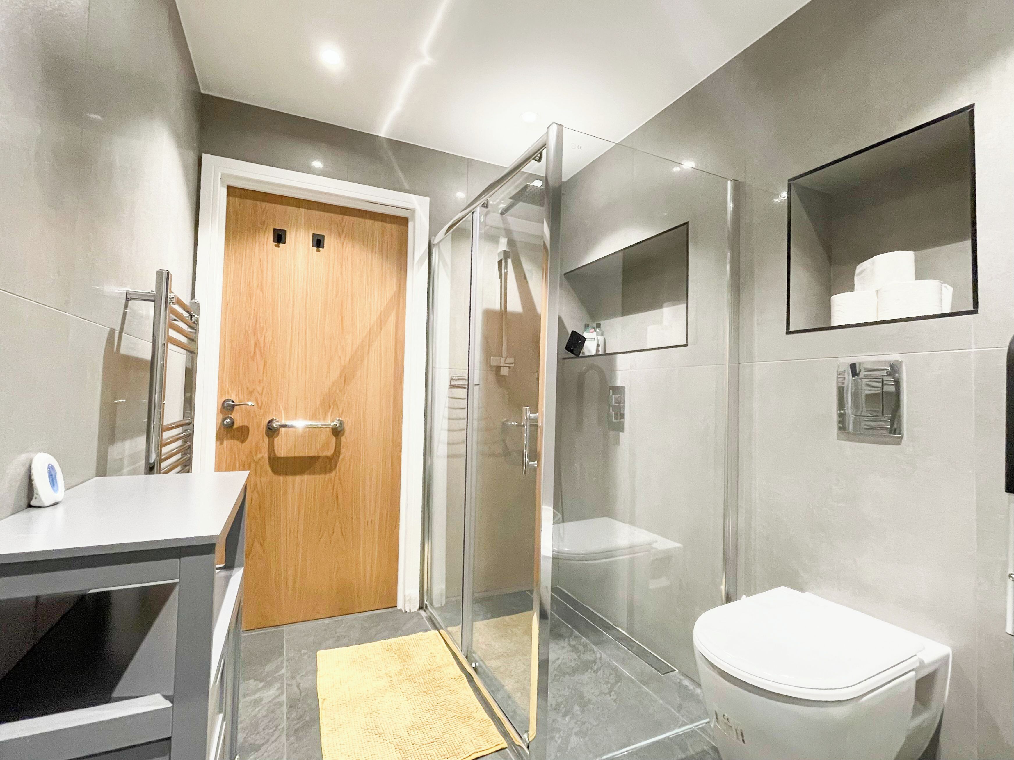 Une salle de bain moderne avec une douche à parois de verre, une porte en bois, des toilettes blanches et un petit miroir au mur.