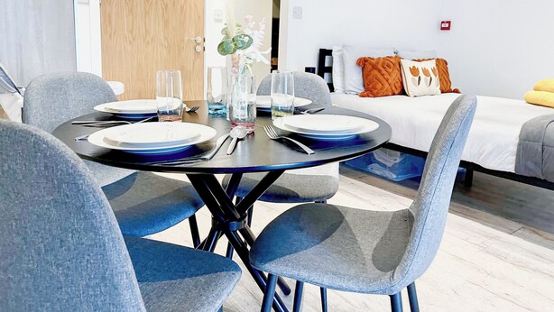 A modern dining area with a round table, grey upholstered chairs, and a white wall with a wooden door in the background.