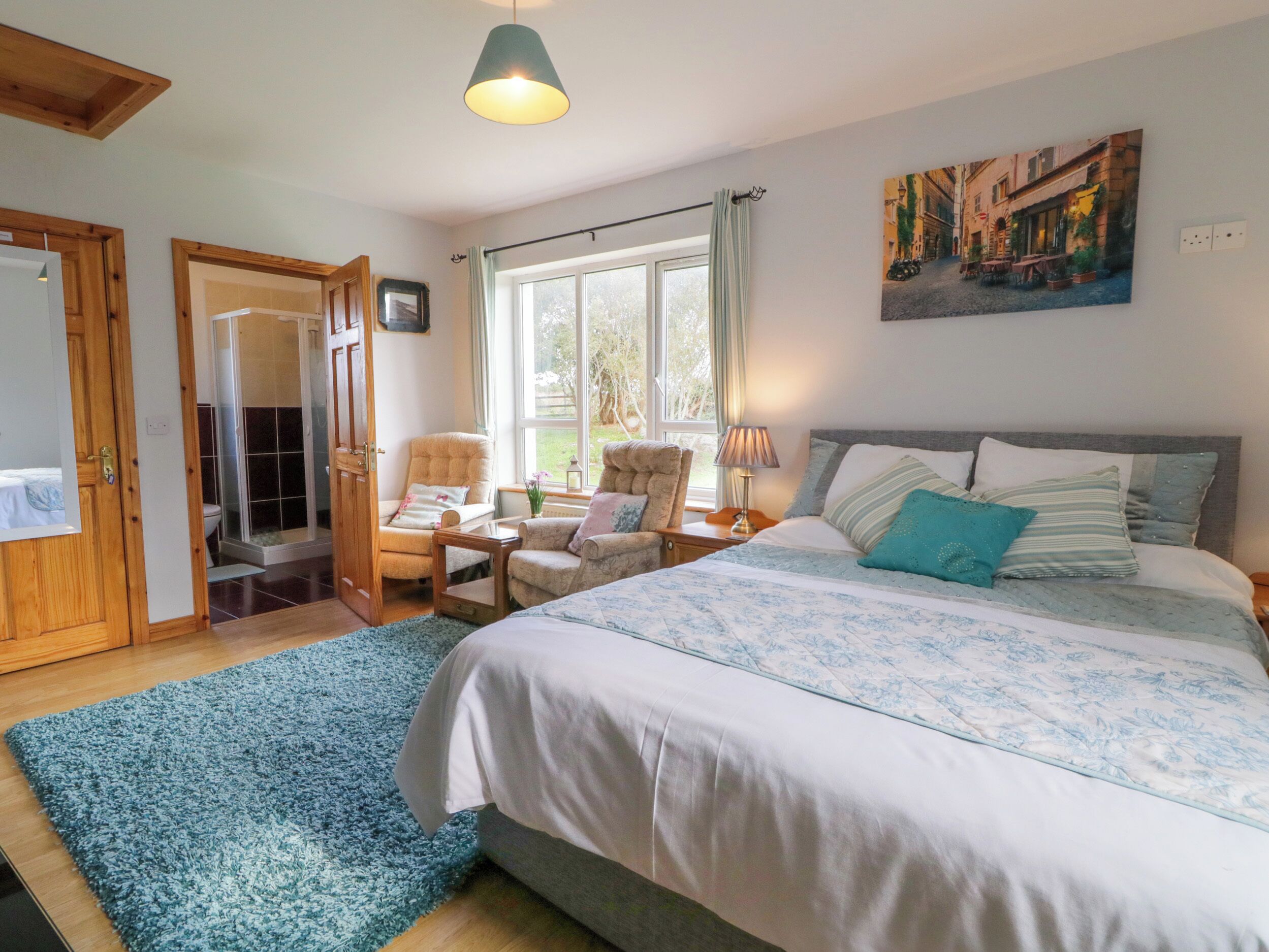 A bedroom with a large bed, two armchairs, a window with curtains, a painting on the wall, and a wooden door.