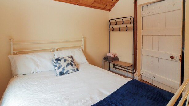 A bedroom with a wooden ceiling, a white bed with a blue and white pillow, a blue blanket, and a metal shelf with towels. - SUSIE'S COTTAGE, pet friendly in Beaufort, County Kerry (KILLARNEY)