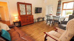 A living room with a brown leather sofa, a wooden cabinet with glass doors, a dining table with chairs, and a television mounted on the wall. - SUSIE'S COTTAGE, pet friendly in Beaufort, County Kerry (KILLARNEY)