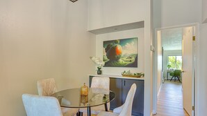 Zona pranzo con tavolo in vetro e sedie bianche, un quadro sopra il tavolo e una cucina sullo sfondo.