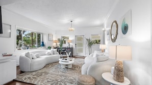 A spacious living room with a large sofa, a coffee table, and a rug. There is a dining area with a table and chairs, and a kitchen in the background. - NEW! Historic Estate-Heated Pool - Two Guest Houses - Sleep16 - NEAR EVERYTHING (West Palm Beach)