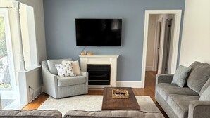 Ein Wohnzimmer mit grauem Sofa, einem grauen Sessel, einem weißen Kamin, einem Flachbildfernseher und einem Holztisch.