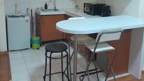 A compact kitchen with a white countertop, a sink, a microwave, and a small refrigerator. There is a folding stool and a green trash bin. - Maytower Service Studio Apartment (Kuala Lumpur)