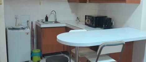 A compact kitchen with a white countertop, a sink, a microwave, and a small refrigerator. There is a folding stool and a green trash bin.