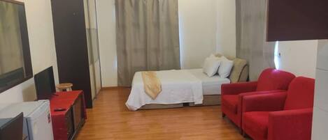 A compact room with a bed, a red chair, a small table, a gas stove, a ceiling fan, and a window with curtains.