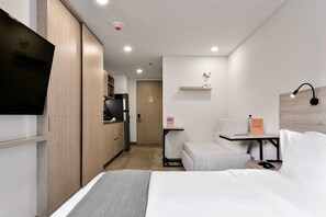 A modern hotel room with a bed, a small desk, a chair, a TV, and a kitchenette.