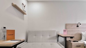 A modern, minimalist room with a grey sofa, a wooden nightstand, a small desk, and a bed with a wooden headboard.