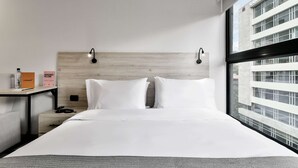 A hotel room with a large bed, a wooden headboard, bedside tables, a nightstand, and a window with a view of a building.