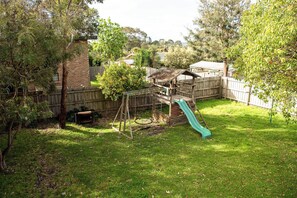 Backyard features a play structure with a slide, surrounded by trees and a grassy lawn.