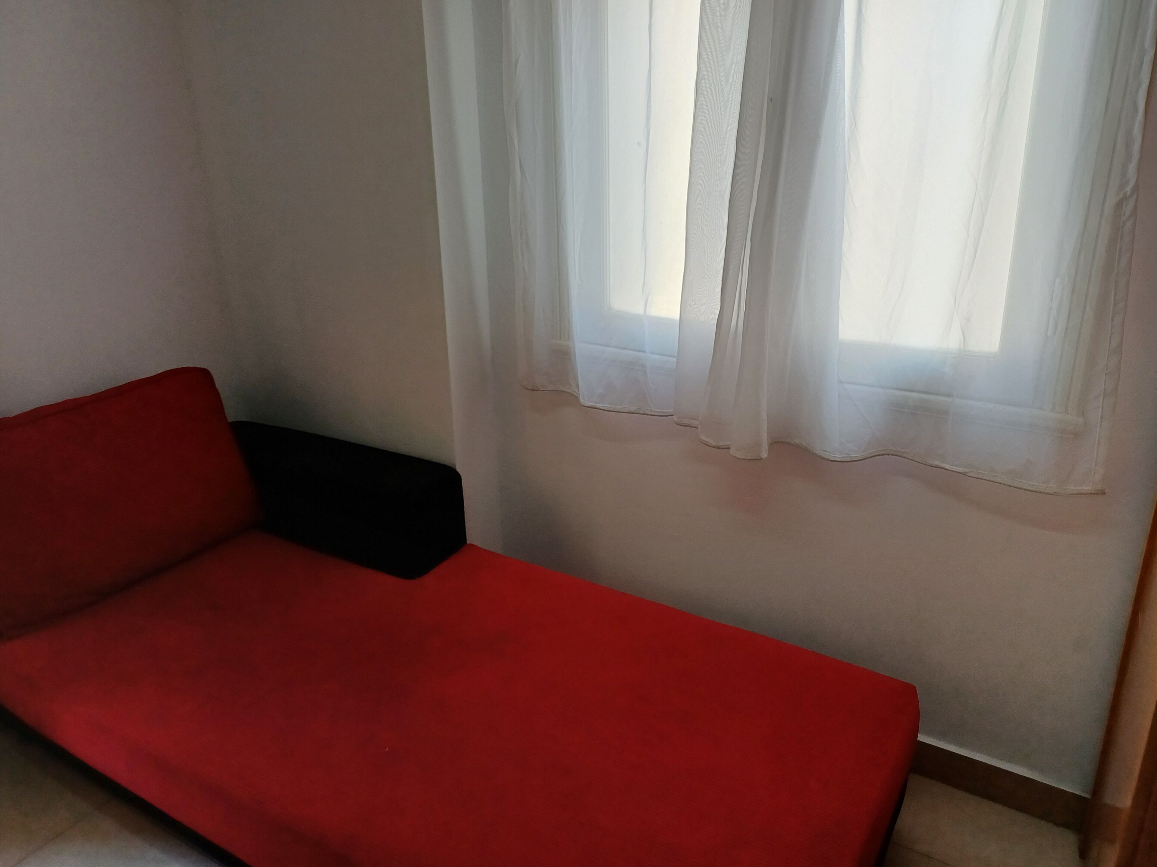 Un sofá rojo en un rincón de la habitación, con una pared blanca y una ventana con cortinas traslúcidas.