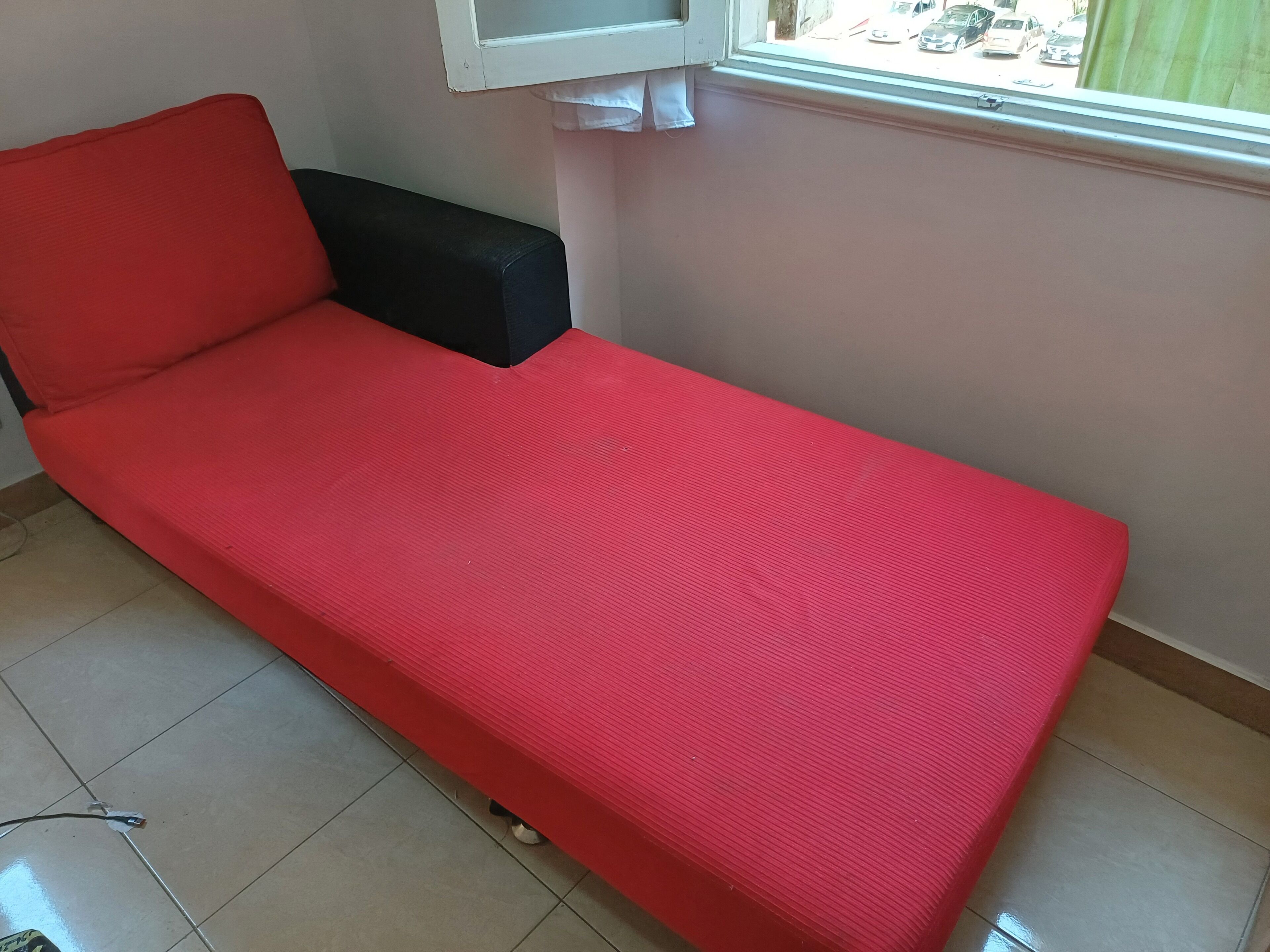 Een kleine eenpersoonskamer met een rode matras, een zwart hoofdeinde en een raam met uitzicht op de straat.