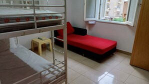 A dormitory room with bunk beds, a small stool, a red sofa, and a window with a view of buildings.
