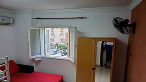 A room with a red sofa, a wooden wardrobe with a mirror, a window with open shutters, and a wall-mounted fan.