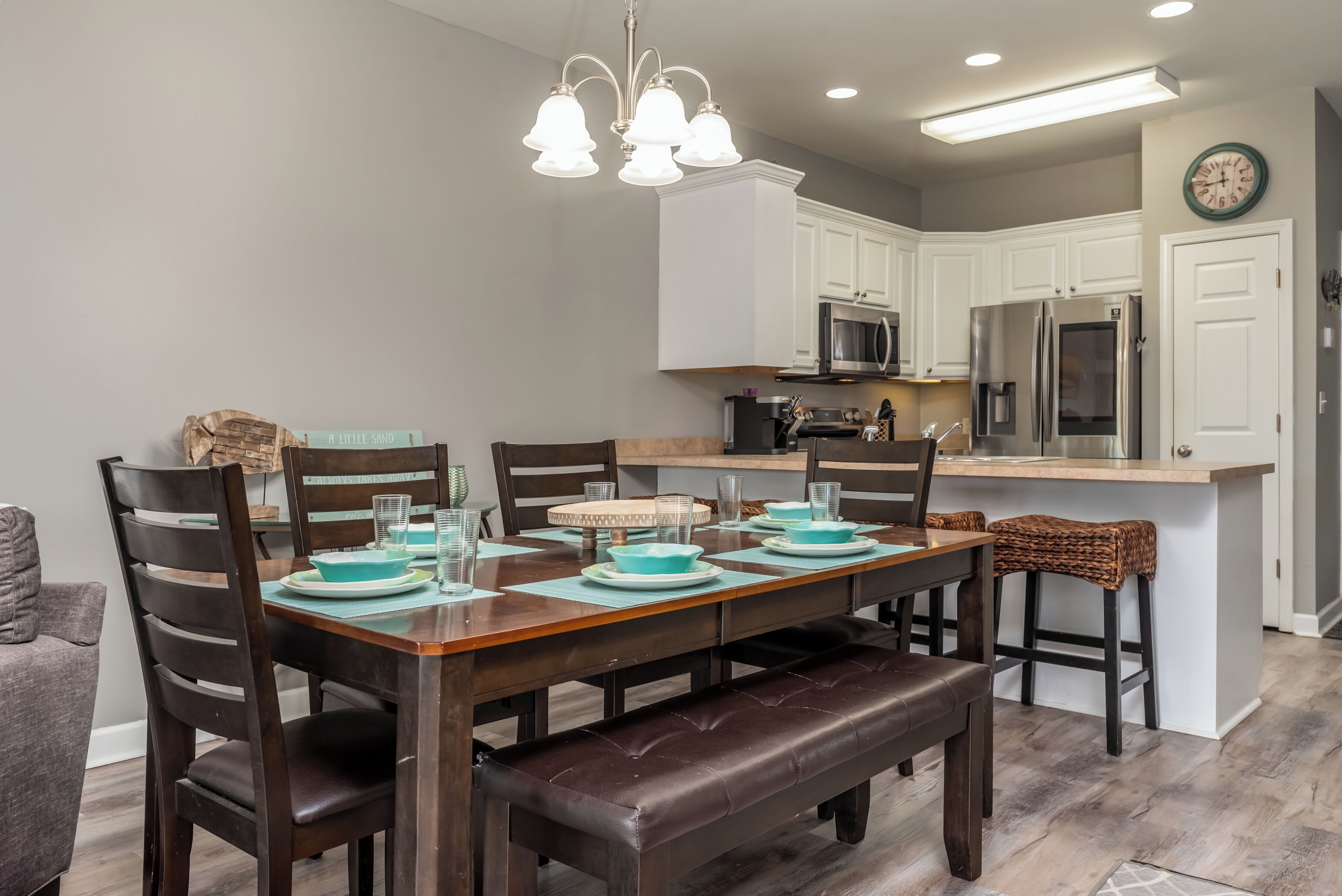 Un área de comedor con una mesa de madera preparada para cuatro personas, rodeada de sillas de madera oscura. Al final de la mesa hay un banco. La habitación cuenta con muebles de cocina blancos, un refrigerador de acero inoxidable y un microondas.