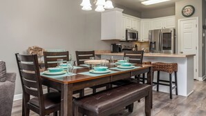 Un área de comedor con una mesa de madera preparada para cuatro personas, rodeada de sillas de madera oscura. Al final de la mesa hay un banco. La habitación cuenta con muebles de cocina blancos, un refrigerador de acero inoxidable y un microondas.