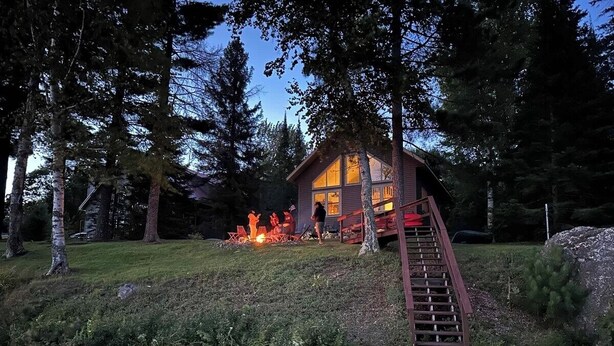 A cabin with a deck and a fire pit, surrounded by trees and a body of water.
