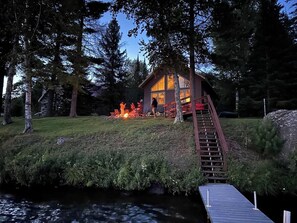 Une cabane dotée d’une terrasse et d’un foyer extérieur, entourée d’arbres et bordée par un plan d’eau.