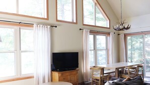 A spacious living room with a large window, a television on a wooden cabinet, a dining table with chairs, and a sofa with a fluffy white throw.