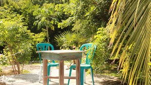 Een houten terras met een tafel en twee stoelen, omgeven door weelderige begroeiing en palmbomen.