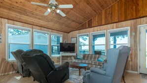 A cozy living room with wooden walls, a ceiling fan, a flat-screen TV, and a large window overlooking a backyard. - Happy Lee Ever After - 1424 Cadillac Avenue 2 Bedroom Home (Dauphin Island)