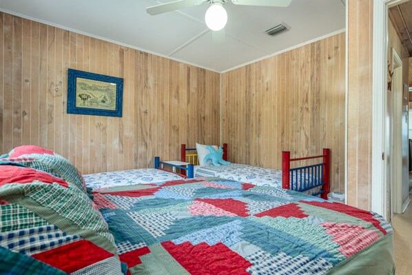 A room with wooden panel walls, two beds with blue and red headboards, a ceiling fan, and a framed picture on the wall.