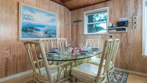 A dining area with a glass table surrounded by wicker chairs, a painting on the wall, and a window with a view of trees. - Happy Lee Ever After - 1424 Cadillac Avenue 2 Bedroom Home (Dauphin Island)
