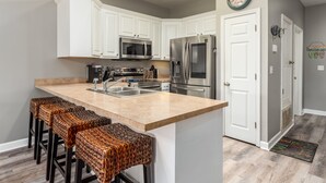 A kitchen with white cabinets, stainless steel appliances, and a bar area with woven stools. - Bayou Heron #2b - 1514 Cadillac 2 Bedroom Condo (Dauphin Island)