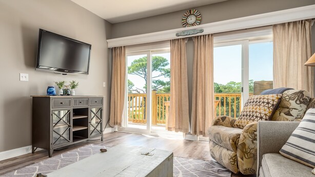 A living room with a flat-screen TV, a cabinet, a sofa, and a sliding glass door to a balcony. - Bayou Heron #2b - 1514 Cadillac 2 Bedroom Condo (Dauphin Island)