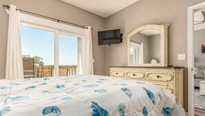 A bedroom with a bed, a dresser, a television, and a large window. - Bayou Heron #2b - 1514 Cadillac 2 Bedroom Condo (Dauphin Island)