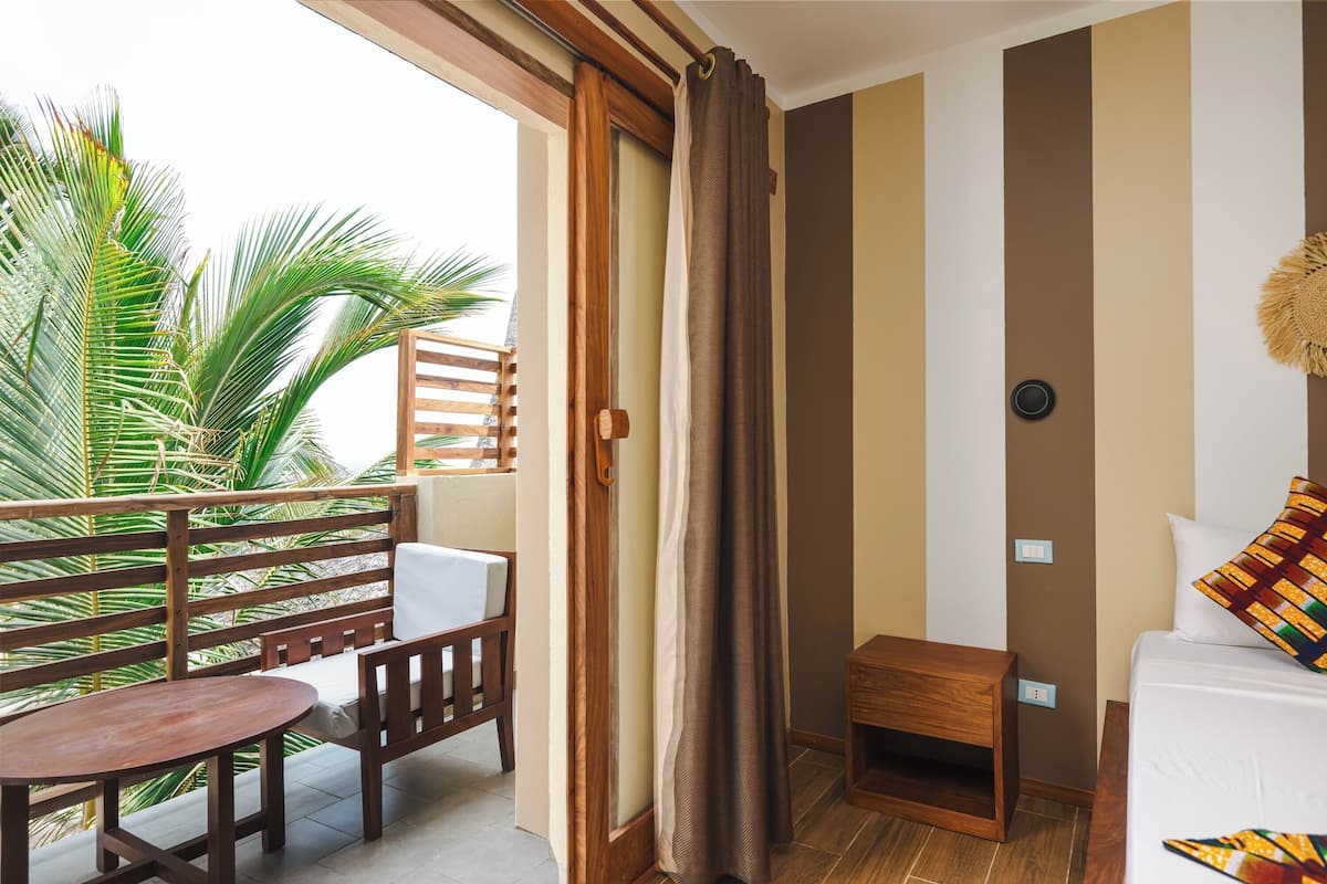 A balcony with a wooden table and chairs, a small wooden nightstand, a bed with a colorful pillow, and a view of palm trees.