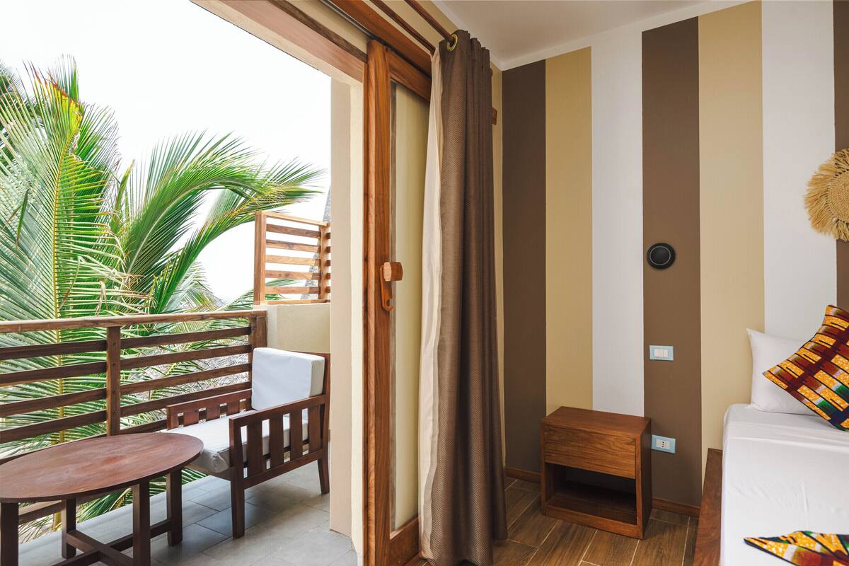A balcony with a wooden table and chairs, a small wooden nightstand, a bed with a colorful pillow, and a view of palm trees.